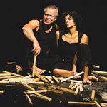 Zwei Personen in schwarzer Kleidung sitzen auf dem Boden, umgeben von verstreuten Holzstäben.