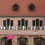 Personengruppe in weißer und beiger Kleidung steht auf einem Balkon vor einem historischen Sandsteingebäude mit rotem Ziegeldach. Im Vordergrund weht eine pinke Fahne vor Rundbögen im Erdgeschoss.