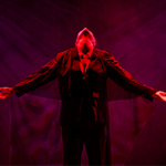 Eine Person breitet die Arme aus und hat den Kopf in den Nacken gelegt um nach oben zu schauen. Sie wird von roten Licht angestrahlt.