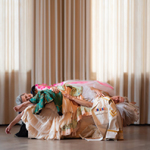 Zwei Tänzerinnen in farbigen Kostümen liegen eng aneinander gelehnt auf einer Couch vor hellen Vorhängen.