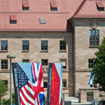 Fassadenansicht eines historischen Sandsteinbaus mit mehreren Fenstern. Vor dem Gebäude stehen drei Fahnenbanner mit den Flaggen der USA, Großbritanniens und der Sowjetunion sowie der Aufschrift „Memorium“. Rechts oben prangt ein blaues Schild mit der weißen Aufschrift „Eintritt frei“.
