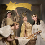 Vier Personen in Kostümen vor dunklem Hintergrund: ein Mann mit Schäferstock und heller Kappe hält einen großen goldenen Stern, daneben drei Menschen in goldenen und weißen Gewändern mit Kronen. Vor ihnen steht ein Korb mit Tuch und einer hölzernen Leier darin.