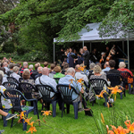 Publikum auf Gartenstühlen verfolgt ein klassisches Konzert unter einem weißen Pavillon in einem üppig grünen Park.