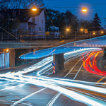 Lichtspuren in Weiß, Blau und Rot schlängeln sich durch eine nächtliche Stadtlandschaft. Eine Brücke überspannt die Straße, während Straßenlaternen ein warmes Licht auf die nasse Fahrbahn werfen. Im Hintergrund sind Wohnhäuser und Bäume zu sehen.