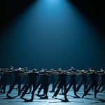 Gruppe von Tänzerinnen und Tänzern in dunkler Kleidung steht im Kreis, synchron mit ausgestreckten Armen zur Seite, unter einem fokussierten, dramatischen Lichtkegel auf dunkler Bühne.