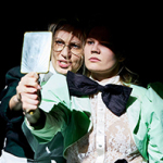 Zwei Personen in dramatischer Beleuchtung, eng beieinander. Die vordere Person trägt ein mintgrünes Jackett, eine transparente Spitzenbluse und eine übergroße schwarze Schleife und hält einen Handspiegel. Neben ihr eine Person mit Brille, die spricht.