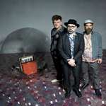 Drei Männer stehen auf einem Boden aus transparenten Schallplatten, im Hintergrund eine große durchsichtige Kugel mit einem kleinen roten Keyboard und weiteren Instrumenten darin.