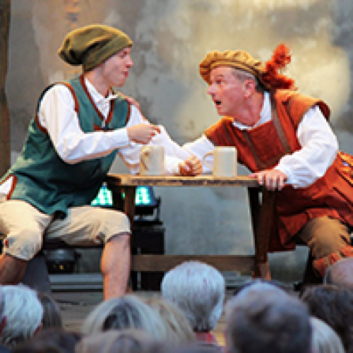 Zwei Männer in mittelalterlicher Kleidung sitzen an einem Holztisch auf einer Bühne im Freien, vor einem Publikum. Einer gestikuliert lebhaft, während der andere zuhört. © Hans-Sachs-Spielgruppe der Stadt Nürnberg