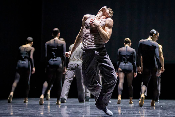 Tänzer in Bewegung auf Bühne, vorn ein Mann mit weißem Tanktop und schwarzer Hose, hinten mehrere Tänzer in schwarzer Kleidung mit Rücken zum Betrachter.