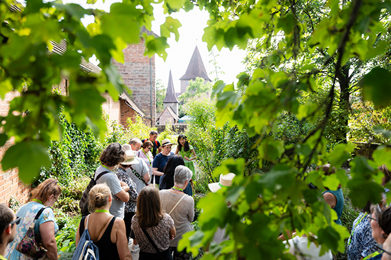 Eine Gruppe von Menschen steht in einem schmalen, grünen Garten zwischen Backsteinmauern und dichtem Laub, im Hintergrund sind spitze Türme und Teile eines historischen Gebäudes zu sehen.