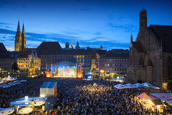 Große Menschenmenge vor beleuchteter Bühne auf dem Hauptmarkt in Nürnberg bei Abenddämmerung mit der Frauenkirche rechts und der Sebalduskirche im Hintergrund.