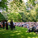 Große Gruppe von Menschen sitzt auf Stühlen im Freien auf einer Wiese vor einer kleinen Bühne mit Musikern, umgeben von Bäumen
