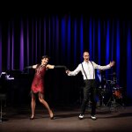 Ein Paar tanzt auf einer Bühne vor einem Orchester mit Klavier, Schlagzeug und Cello vor einem dunkelblauen Bühnenvorhang