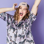 Junge Frau mit langem Haar, welche ein ein helles Hemd mit Zebramuster und eine Schlafmaske mit Tiergesicht trägt, reckt sich.