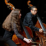 Musikerinnen und Musiker sitzen nebeneinander und spielen Kontrabass auf einer Bühne