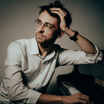 Mann mit Brille und weißem Hemd sitzt nachdenklich an einem Klavier und fasst sich an den Kopf