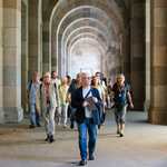 Eine Menschengruppe geht durch einen von steinernen Rundbögen gesäumten Gang