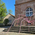 Große, rosa Metallskulptur einer Spinne mit langen, dünnen Beinen vor einem Sandsteingebäude