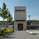 Eingangsbereich des Museums Industriekultur in Nürnberg mit Betonfassade, Schriftzug »MUSEUM«, umgeben von Bäumen und blühenden Sträuchern