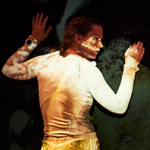 Person mit hochgestecktem Haar und weißem Langarmshirt hebt beide Hände vor einer dunklen Wand mit unscharfen Projektionen