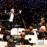 Dirigent mit erhobenen Armen vor Orchester und Publikum mit vielen Lichtern im Hintergrund