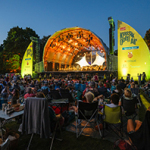 Open-Air-Konzertbühne mit beleuchtetem Dach und Publikum auf einer Wiese bei Dämmerung