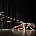 Person in sportlicher Kleidung zieht knieend auf einer Bühne ein Seil mit einem Stapel von Stühlen