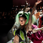 Mehrere Tänzerinnen in bunten Kostümen mit erhobenen Armen auf einer Bühne vor dunklem Publikumshintergrund