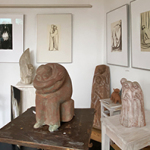 Skulpturenausstellung mit mehreren abstrakten Figuren aus Ton auf Tischen und Podesten in einem weißen Raum mit gerahmten Zeichnungen an den Wänden