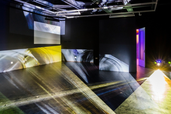 Dunkler Raum mit mehreren projizierten Licht- und Bildflächen sowie offenstehender Tür mit violettem Lichtschein