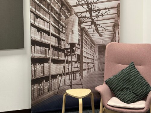 Ein Lesesessel und Beistelltisch stehen vor einer Fototapete, auf der ein Mann auf einer Leiter an einem hohen, gefüllten Bücherregal stehend zu sehen ist.