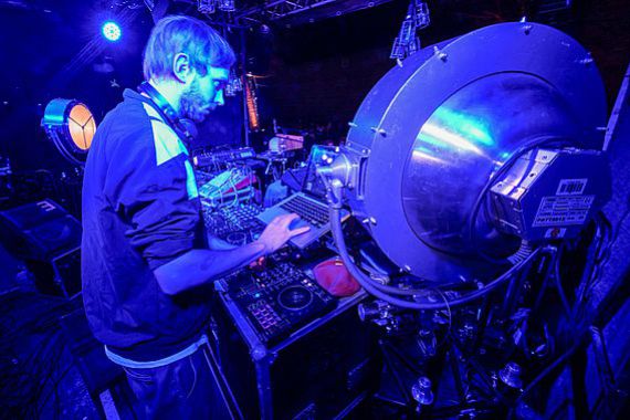 Ein DJ von We All Die Worse steht auf der blau beleuchteten Bühne in Mitte von Scheinwerfern, Computern und Synthesizern.
