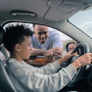 Ein junger Mann sitzt im Auto am Lenkrad. Ein älterer Mann steht am Fenster und erklärt etwas.
