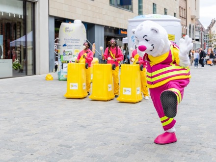 Foto: Der Trenn-Bär in der Fuß·gänger·zone. Links danaben trommeln 3 Personen auf umgedrehten Gelben Tonnen., Bild © Maik Ludwig / ADvantage Production / Initiative „Mülltrennung wirkt“