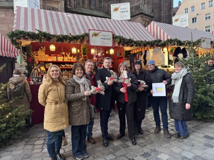 Foto von 8 Personen mit dem neuen Kuschel·hasen Albrecht. Im Hintergrund ist die original-regional-Bude vom Christ·kindlesmarkt zu sehen., Bild © Kerstin Stübs / Stadt Nürnberg