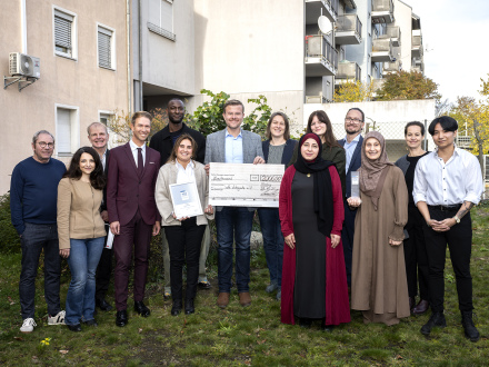 Foto von 14 Personen auf einer Wiese vor einem Haus. Die Personen gehören zum Projekt Digi iQ und zum Verein We Integrate e.V. In der Mitte steht der Ober·bürgermeister Marcus König mit einem großen Scheck aus Papier in der Hand. Rechts neben ihm steht die Sozial·referentin von Nürnberg.
