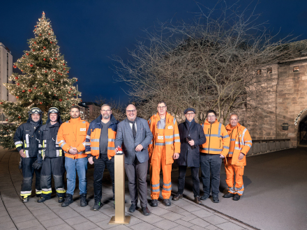 Foto vom großen Weihnachts·baum auf dem Bahnhofs·platz. Vor dem Weihnachts·baum steht Christian Vogel mit dem Team vom Weihnachts·baum.