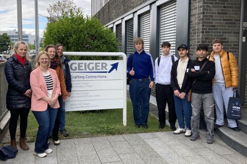 Betriebsbesichtigung bei der Geiger Gruppe. Jugendliche Teilnehmende von SCHLAU, Mitarbeitende von SCHLAU und Mitarbeiterin von der Geiger Gruppe stehen vor dem Firmenschild.