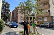 Karsten Neumann mit einem Kunstobjekt und Baummanager Florian Demling mit der Spendenbanderole von Bethang stehen vor dem neuen Baum in der Kreutzerstraße