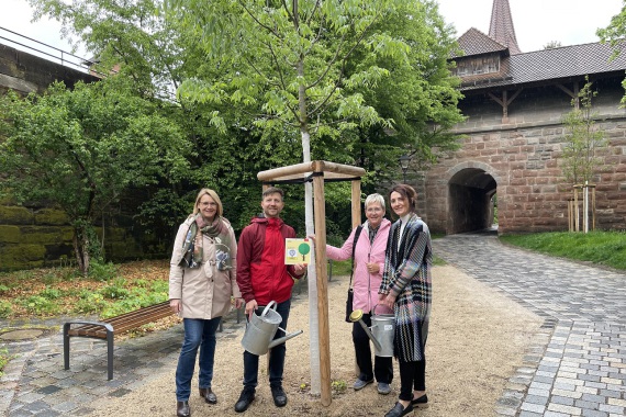 Drei Mitglieder und Präsidentinnen des SI Club Nürnberg stehen mit dem Stadtbaum-Manager am Spendenbaum. Sie halten zwei Gießkannen aus Metall und das Spendenschild. Im Hintergrund das Hallertor.