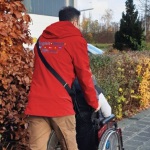 Ein Mann mit roter Jacke mit der Aufschrift Begleit-Dienst schiebt eine Person im Rollstuhl