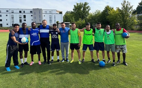 Eine Gruppe von elf jungen Männern steht auf einem Fußballfeld in einer Reihe und lächelt in die Kamera. Sie tragen Sportkleidung; die linke Mannschaft trägt überwiegend blaue Trikots, die rechte Mannschaft grüne Leibchen. Zwei Spieler halten jeweils einen Ball. Im Hintergrund stehen moderne Wohngebäude und Bäume.