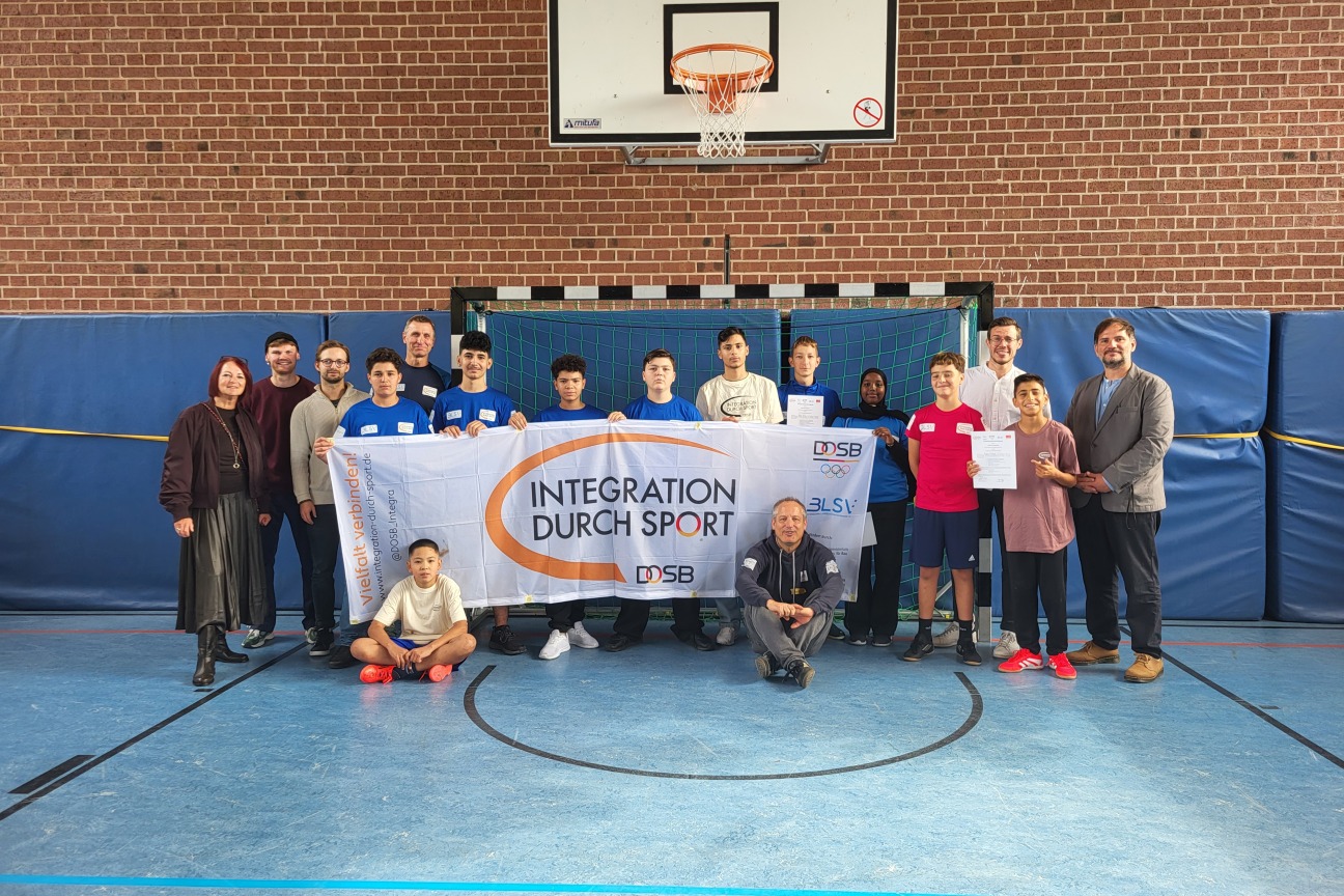 Das Bild zeigt eine Gruppe von Jugendlichen und Erwachsenen in einer Sporthalle. Sie stehen zusammen und halten ein Banner mit der Aufschrift „INTEGRATION DURCH SPORT“, das von einem der Jugendlichen gehalten wird. Einige der Teilnehmer halten Zertifikate in den Händen. Die Gruppe besteht aus Jungen unterschiedlichen Alters und Herkunft, sowie einigen Erwachsenen, darunter eine Frau und ein Mann, die beide am linken und rechten Rand des Bildes stehen. Der Hintergrund zeigt ein Fußballtor und die typischen Merkmale einer Sporthalle. Die Atmosphäre wirkt feierlich, und es scheint, als ob ein Event oder eine Auszeichnung im Rahmen des Projekts „Integration durch Sport“ stattfindet.