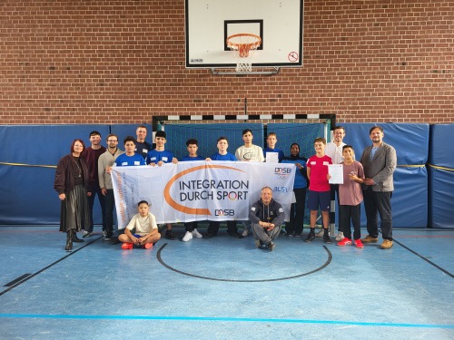 Das Bild zeigt eine Gruppe von Jugendlichen und Erwachsenen in einer Sporthalle. Sie stehen zusammen und halten ein Banner mit der Aufschrift „INTEGRATION DURCH SPORT“, das von einem der Jugendlichen gehalten wird. Einige der Teilnehmer halten Zertifikate in den Händen. Die Gruppe besteht aus Jungen unterschiedlichen Alters und Herkunft, sowie einigen Erwachsenen, darunter eine Frau und ein Mann, die beide am linken und rechten Rand des Bildes stehen. Der Hintergrund zeigt ein Fußballtor und die typischen Merkmale einer Sporthalle. Die Atmosphäre wirkt feierlich, und es scheint, als ob ein Event oder eine Auszeichnung im Rahmen des Projekts „Integration durch Sport“ stattfindet.