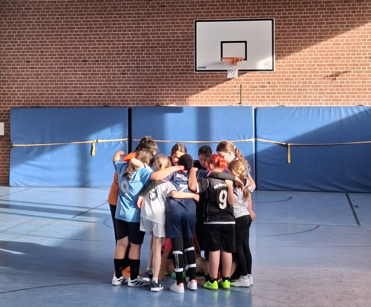 Eine Gruppe von Kindern steht eng im Kreis in einer Sporthalle, die Arme umeinander gelegt, unter einem Basketballkorb vor einer Backsteinwand., Bild © Leon Ackermann