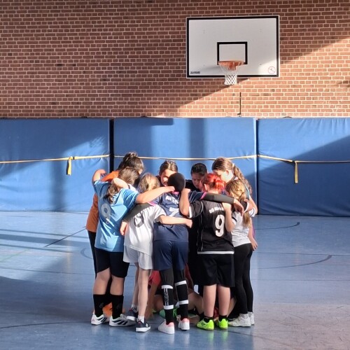 Eine Gruppe von Kindern steht eng im Kreis in einer Sporthalle, die Arme umeinander gelegt, unter einem Basketballkorb vor einer Backsteinwand.