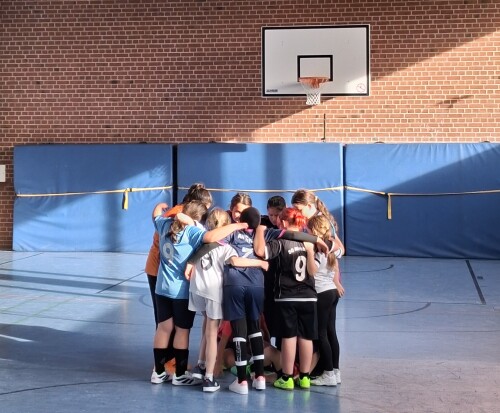 Eine Gruppe von Kindern steht eng im Kreis in einer Sporthalle, die Arme umeinander gelegt, unter einem Basketballkorb vor einer Backsteinwand.