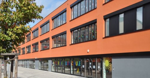 Modernes Schulgebäude der Kopernikus Schule Nürnberg mit oranger Fassade und großen Fenstern, im Erdgeschoss bunte Buchstaben und Sonnenmotiv an den Fenstern.