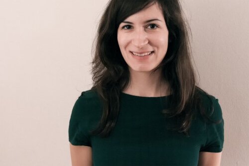 Porträt einer lächelnden Frau mit schulterlangem dunklem Haar, die ein dunkelgrünes Oberteil trägt und vor einer hellen, neutralen Wand steht. © Julia Stegerer