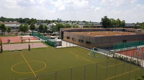 Außenansicht eines Schul- und Sportgeländes mit Fußballfeld im Vordergrund, mehreren Tennisplätzen und einem flachen, modernen Gebäude mit Holzfassade unter leicht bewölktem Himmel. © Horst Heil